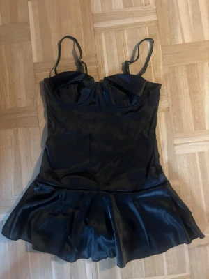Svart satin kortklänning med kupor - Kort svart klänning i satin med smala axelband och markerade kupor. Klänningen har en lätt utsvängd kjol och figurnära passform upptill för en trendig och elegant siluett.