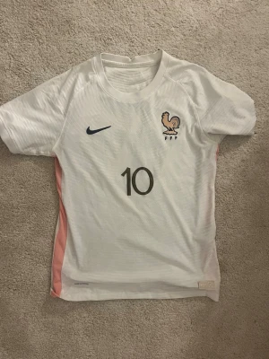 Vit Frankrike Nike fotbollströja #10 - Snygg vit fotbollströja från Nike med Frankrikes landslagsemblem och nummer 10 på bröstet. Tröjan har blå Nike-logga, broderad tupp och FFF-detaljer. Rosa paneler på sidorna och tillverkad i lätt, andningsbart material perfekt för träning eller match.