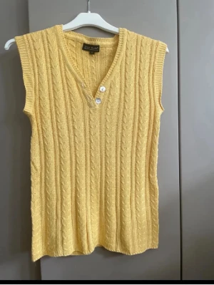 Gul stickad topp från Vero Moda - Snygg gul stickad topp från Vero Moda med kabelstickat mönster och v-ringning. Västen har två knappar framtill och är ärmlös, perfekt att styla över en skjorta eller t-shirt. Materialet är en mjuk mix av viskos och bomull.