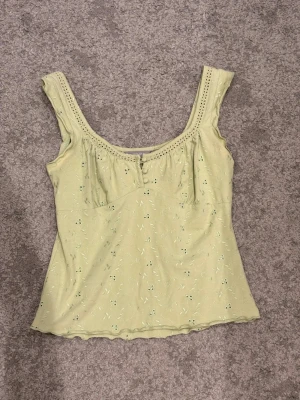Ljusgrön blommig linnetopp - Supersöt ljusgrön linnetopp med små blommiga broderier och breda axelband. Toppen har en lätt vågig kant nertill och dekorativ hålbrodyr vid ringningen. Perfekt för varma dagar och enkel att styla med jeans eller kjol.