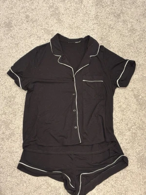 Svart pyjamasset med vita detaljer - Snygg svart pyjamas i två delar med kortärmad skjorta och shorts. Setet har vita kontrastsömmar längs kanterna och knappar framtill på toppen. Skjortan har en bröstficka och klassisk krage. Perfekt för mysiga kvällar hemma.