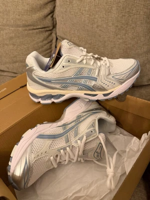 Vita och silver Asics sneakers - Snygga vita sneakers från Asics med detaljer i silver och ljusblått. Skorna har en sportig design med meshpaneler för bra ventilation och en rejäl sula med bra grepp. Perfekta för dig som gillar en clean och modern look.