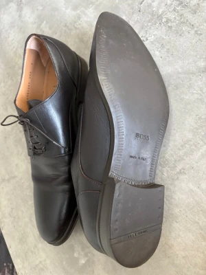 Svarta skinnskor från Hugo Boss - Klassiska svarta skinnskor från Hugo Boss, tillverkade i Italien. Skorna har snörning, rund tå och en elegant, tidlös design. Perfekta för dig som vill ha en stilren look med premiumkänsla.