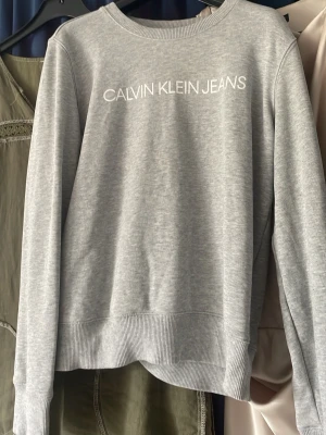Grå sweatshirt från Calvin Klein Jeans - Snygg ljusgrå sweatshirt från Calvin Klein Jeans med vit logga tryckt över bröstet. Tröjan har rund halsringning, ribbade muddar vid ärmslut och nederkant samt en avslappnad passform. Perfekt till jeans eller mjukisbyxor.