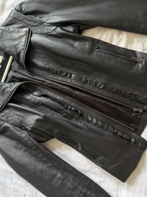 Svart skinnjacka med dragkedjor - Snygg svart skinnjacka med flera dragkedjor framtill och coola detaljer i form av dekorativa sömmar och volangliknande kant. Jackan har en klassisk krage och raka ärmar. Perfekt för dig som vill ha en edgy och stilren look.