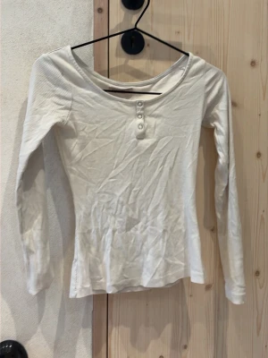 Vit långärmad topp med knappar - Snygg vit långärmad topp med rund halsringning och tre dekorativa knappar framtill. Modellen är figurnära och passar perfekt till jeans eller kjol. Enkel och stilren design som funkar till det mesta.