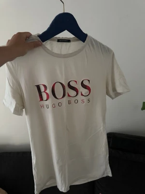 Vit Hugo Boss t-shirt med logga - Snygg vit t-shirt från Hugo Boss med stor BOSS-logga i rött och svart framtill. Klassisk rund halsringning och korta ärmar. Perfekt till jeans eller shorts för en clean och stilren look.