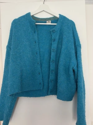Blå fluffig kofta från Urban Outfitters - Mysig blå kofta i fluffigt material från Urban Outfitters. Koftan har rund halsringning, långa ärmar och knäppning framtill med flera knappar. 