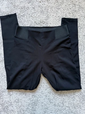 Svarta leggings med bred midjeresår - Säljer ett par svarta leggings med hög midja och bred resår som ger en snygg och bekväm passform. Materialet är stretchigt och mjukt, perfekt för en avslappnad look. Inga synliga fickor eller detaljer, bara clean och stilren design.