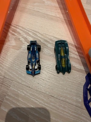 Hot Wheels bilbana med två bilar - Säljer en Hot Wheels bilbana med två racingbilar i metall, en blå och en grön. Banan har två orange spår, startmekanismer och en looping. Perfekt för tävlingar hemma och för dig som gillar fartfyllda leksaker.