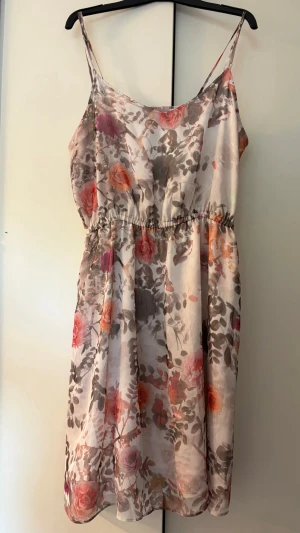 Blommig sommarklänning med smala axelband - Söt blommig klänning med smala axelband och elastisk midja. Klänningen har en ljus bas med rosa, orange och lila blommor samt gröna blad. Tunn och luftig, perfekt för varma dagar. Längden är ovanför knät och materialet känns som polyester. Från Anna Field, storlek 44.
