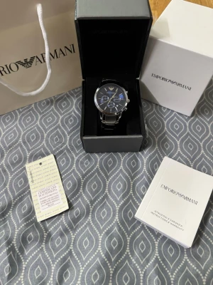 Emporio Armani silverfärgad klocka blå - Stilren klocka från Emporio Armani med blå urtavla och silverfärgat armband i metall. Klockan har kronograf-funktion, datumvisning och klassisk rund form. Levereras i originalask med certifikat och manual.