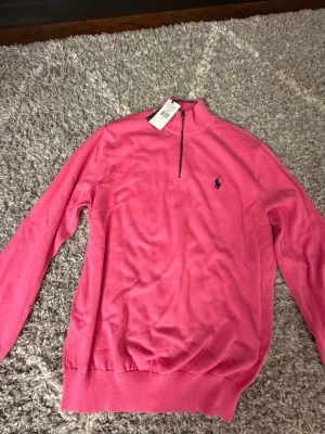 Rosa stickad tröja från Polo Golf - Snygg rosa stickad tröja från Polo Golf Ralph Lauren med halv dragkedja och broderad logga på bröstet. Tröjan har ribbade muddar och krage, perfekt för en sportig och avslappnad look. Materialet känns mjukt och skönt mot huden.