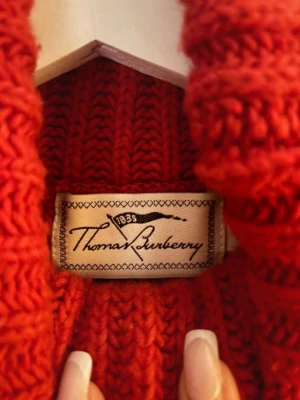 Röd polotröja från Thomas Burberry - Säljer en chunky röd stickad polotröja från Thomas Burberry. Tröjan har grov ribbstickning, hög krage, ficka framtill och dekorativa knappar på sidan. Perfekt för kalla dagar och riktigt snygg till jeans.