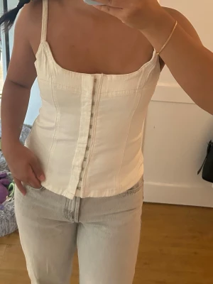 Vit jeans topp - Säljer en vit topp med smala axelband och knappar framtill. Toppen har en figurnära passform och markerade sömmar som ger en snygg siluett. Perfekt att styla med jeans eller kjol för en trendig look.