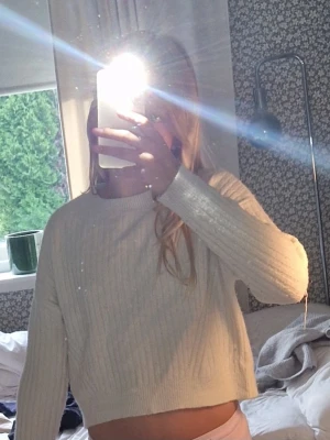 Vit ribbstickad cropped tröja - Säljer en vit ribbstickad cropped tröja med rund hals och långa ärmar. Tröjan har en enkel och stilren design med en liten brun detalj nertill. Perfekt att matcha med höga jeans eller kjol. Pris kan ändras
