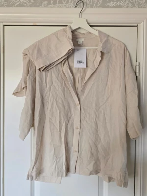 Beige linneset från H&M - Luftig och avslappnad beige skjorta och shorts från H&M i linne. Skjortan har klassisk krage, knappar framtill och trekvartsärm. Perfekt för varmare dagar och enkel att styla med olika outfits. Shortsen har resår i bak.