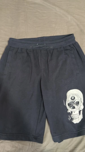 Svarta shorts med dödskalletryck - Säljer ett par svarta shorts med snörning i midjan och ett stort vitt dödskalletryck på ena benet. Shortsen har sidofickor och är tillverkade i mjukt bomullsmaterial. Perfekta för en avslappnad och edgy stil.