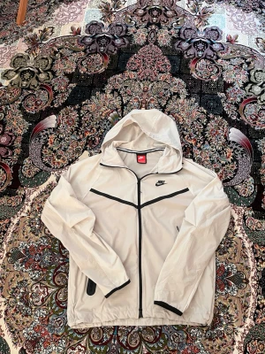 Vit vindjacka från Nike med huva - Snygg vit vindjacka från Nike med svarta detaljer och dragkedja framtill. Jackan har huva, två sidofickor och en sportig design med svarta linjer över bröstet. Perfekt för blåsiga dagar och enkel att matcha till olika outfits.