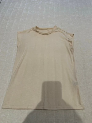 Beige ärmlös topp i bomull - En enkel och stilren beige topp utan ärmar, tillverkad i mjuk bomull. Toppen har rund halsringning och rak passform, perfekt att matcha med jeans eller kjol för en clean look.