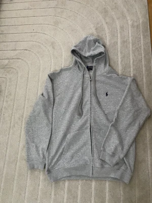 Grå zip hoodie från Polo Ralph Lauren - Snygg grå hoodie med dragkedja från Polo Ralph Lauren. Klassisk design med huva och dragsnören, två fickor framtill och den ikoniska lilla loggan broderad på bröstet. Tillverkad i mjukt bomullsmaterial som är skönt att ha på sig.