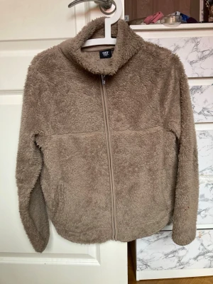 Beige teddyjacka från 157 - Supermjuk teddyjacka i beige från 157 med dragkedja framtill och hög krage. Jackan har lång ärm och är tillverkad i fluffigt teddy-material som ger en mysig och trendig look. Perfekt för lager-på-lager och chill dagar.