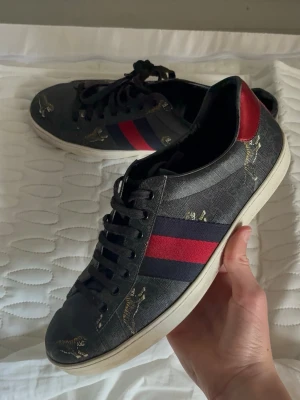 Gucci sneakers med tigerprint - Snygga Gucci sneakers i svart läder med klassiska röda och blå ränder på sidan samt tigerprint över hela skon. Skorna har vita sulor, svarta skosnören och kontrasterande hälflikar i rött och blått. Perfekta för dig som vill sticka ut med exklusiv stil.