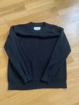 Svart stickad tröja från maison margiela - Klassisk svart stickad tröja från maison margiela med rund halsringning och ribbade muddar. Tröjan har en rak passform och är tillverkad i ett mjukt, grovstickat material som passar perfekt till lager-på-lager-stil.