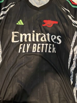Arsenal svart matchtröja Saka #7 - Snygg svart Arsenal matchtröja från Adidas med Saka #7 på ryggen. Tröjan har vita detaljer, grönt Adidas-märke och rött Arsenal-emblem. Tillverkad i lätt och ventilerande polyester, perfekt för fotboll eller träning. ENDAST UPPHÄMTNING I STOCKHOLM!!