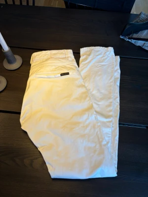 Vita chinos från Nudie Jeans - Säljer ett par vita chinos från Nudie Jeans med slim fit passform. Byxorna har klassiska fickor fram och bak, samt diskret logga ovanför bakfickan.  Materialet känns mjukt och följsamt. Pris kan diskuteras!