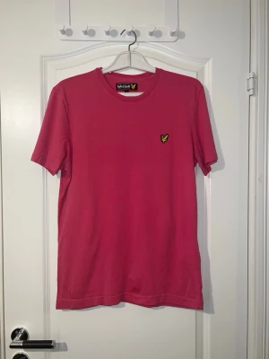 Rosa t-shirt från Lyle & Scott - Snygg rosa t-shirt från Lyle & Scott med klassisk rund hals och det ikoniska gula logomärket broderat på bröstet. Tillverkad i mjuk bomull och har en normal passform. Perfekt för en avslappnad och stilren look. Finns att hämta i Södertälje.