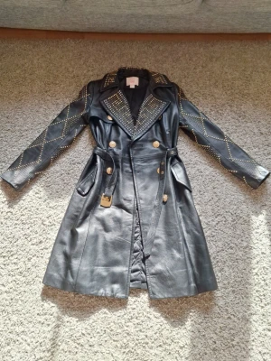 Versace x H&M black leather studded trenchcoat  - Versace x H&M svart trenchcoat med lädernitar i storlek 36. Använd fåtal gånger och skicket är jättebra. Den har två fickor och många detaljer som lädernitarna och gulliga Versace-knappar. Denna jack är sjuk sällsynt nu för tiden och går för ca 4000-5000 kr. Den är supertrendig just nu och passar alla outfits. Skick: 9/10
