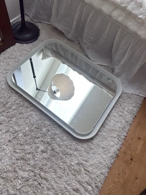 Rektangulär spegel med vit ram - Modern rektangulär spegel med bred vit ram och rundade hörn. Spegeln har en stilren design och är tillverkad av glas och plast. Perfekt som inredningsdetalj i sovrum, hall eller vardagsrum.