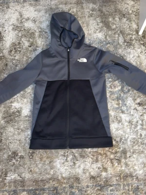 Grå och svart hoodie från The North Face - Snygg hoodie från The North Face i grått och svart med dragkedja framtill och huva. Loggan är tryckt på bröstet och det finns en praktisk ficka med dragkedja på ena ärmen. Perfekt för chill eller streetwear.