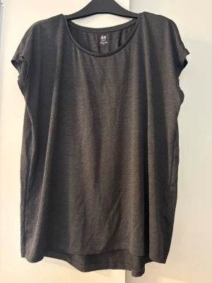 Mörkgrå sporttopp från H&M - En enkel och snygg mörkgrå sporttopp från H&M Sport. T-shirten har rund halsringning, korta ärmar och är gjord i ett mjukt, lätt material som passar perfekt till träning eller chill. Stilren design utan tryck eller mönster.