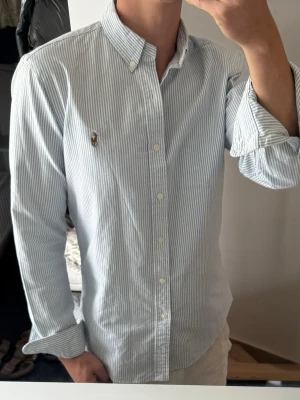 Randig skjorta från Ralph Lauren - Klassisk blå- och vitrandig skjorta från Ralph Lauren med button-down krage och broderad logga på bröstet. Skjortan har långa ärmar och knappar framtill. Perfekt till jeans eller chinos för en clean och stilren look.