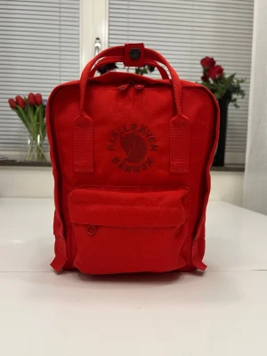 Röd Fjällräven Re-Kånken Mini ryggsäck - Säljer en röd Fjällräven Re-Kånken Mini ryggsäck i slitstark canvas. Klassisk fyrkantig form med dragkedja, framficka och två sidofickor. Justerbara axelband och handtag upptill. Ikonisk broderad logga på framsidan. Knappt använt!!!!                                  Höjd: 29 cm Bredd: 20 cm Djup: 13 cm Volym: 7 l Vikt: 340 g Antal fickor: 4 Sittunderlag ingår: Ja