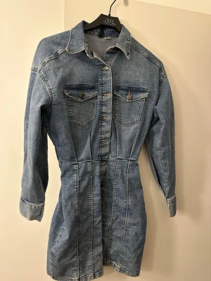 Jeansklänning från DIVIDED - Snygg jeansklänning från DIVIDED i klassisk blå denim. Klänningen har lång ärm, knappar framtill och två bröstfickor med lock. Figursydd modell som ger en cool och avslappnad look. Perfekt att styla med sneakers eller boots.