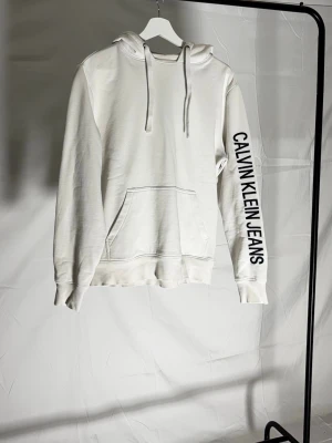 Vit hoodie från Calvin Klein Jeans - Sick 6/10 Skada vid baksida av axel, inga andra defekter.                                                                  Snygg vit hoodie från Calvin Klein Jeans med marinblå logga längs ena ärmen och tryck på huvan. Klassisk känguruficka framtill och justerbar dragsko i huvan. Perfekt för en avslappnad och trendig look.
