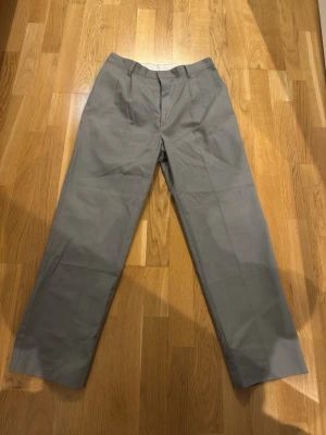 Grå kostymbyxor med raka ben - Snygga grå kostymbyxor med klassisk rak passform och pressveck. Byxorna har hällor för skärp och diskreta fickor både fram och bak. Perfekta för en clean och stilren look. Materialet känns slätt och har en lätt glans.