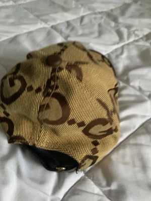 Gucci beige keps med GG-mönster - Säljer en beige keps från Gucci med det klassiska bruna GG-monogrammet över hela kepsen. Kepsen har böjd skärm med brun kant och justerbart band bak. Materialet är canvas med detaljer i läder. Perfekt för dig som gillar exklusiva accessoarer.