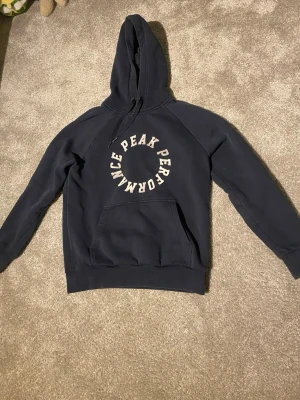 Mörkblå hoodie från Peak Performance - Snygg mörkblå hoodie från Peak Performance med stor vit logga i cirkelform på bröstet. Klassisk känguruficka framtill och justerbar huva med dragsko. Perfekt för en avslappnad och sportig stil.