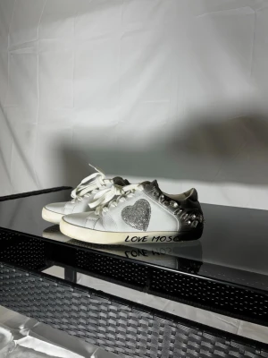 Love Moschino vita sneakers med hjärta - Sick 7/10.                                                                  Snygga vita sneakers från Love Moschino med silverdetaljer, glittrigt hjärta på sidan och nitar vid hälen. Skorna har texten 'LOVE MOSCHINO' på sulan och vita snören. Perfekta för dig som vill sticka ut med coola detaljer och trendig design.