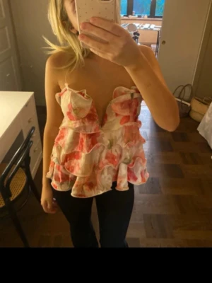 Blommig volang tubtopp i rosa och vitt - Säljer en romantisk axelbandslös tubtopp med blommigt mönster i rosa, vitt och grönt. Toppen har flera lager volanger och en v-ringad skärning framtill. Perfekt för dig som vill sticka ut med en feminin och trendig look.