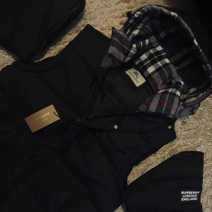 Svart dunväst från Burberry - Snygg svart dunväst från Burberry med avtagbar huva och klassiskt rutigt foder i grått, vitt och rött. Västen har dragkedja och tryckknappar framtill samt två fickor. Perfekt för lager på lager och streetstyle.