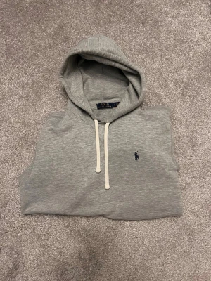 Grå hoodie från Polo Ralph Lauren - Snygg grå hoodie från Polo Ralph Lauren med klassisk huva och vita dragsnören. Ikoniska blå loggan broderad på bröstet. Tillverkad i mjukt bomullsmaterial som känns riktigt skönt mot huden. Perfekt för en chill och stilren look.