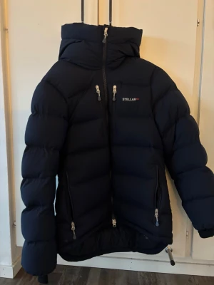 Mörkblå dunjacka från Stellar equipment  - Snygg mörkblå dunjacka från Stellar med stor huva och flera dragkedjeförsedda fickor. Jackan har en puffig design och logga på bröstet samt ryggen. Går både att ha till vardags eller i skidbacken. Perfekt vinterjacka med 800 i fillpower. Jackan är förra säsongens dammodell. I fint skick förutom lite smink fläckar som syns på bilderna! Nypris 5995