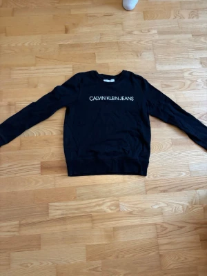 Svart sweatshirt Calvin Klein Jeans - Svart sweatshirt från Calvin Klein Jeans med vit logotyptext på bröstet. Klassisk rund halsringning och långa ärmar. Tillverkad i mjukt bomullsmaterial för en bekväm och avslappnad stil.