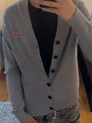 Grå cardigan från Scotch & Soda - Snygg grå cardigan från Scotch & Soda med svart knappstängning och röd broderad logga på bröstet. Tröjan har ribbade muddar och är tillverkad i mjukt material. Den är i väldigt bra skick. Den är en storlek S men jag skulle säga att den är en M. Modellen i bilden är 188cm och väger 74kg. 