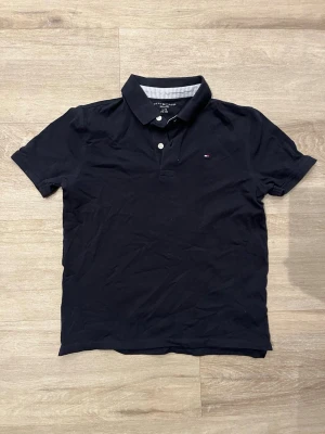 Marinblå piké från Tommy Hilfiger - Snygg marinblå pikétröja från Tommy Hilfiger med klassisk krage och två knappar framtill. Liten broderad logga på bröstet och diskret randigt tyg på insidan av kragen. Tillverkad i mjuk bomull för en bekväm känsla.